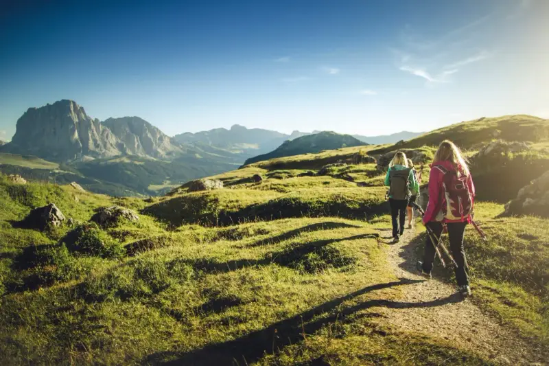 Persone in un percorso di trekking e hiking tra le montagne, con zaini e bastoncini da passeggio, sotto un cielo azzurro.